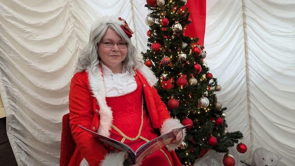 Person in red Santa outfit reading book next to decorated Christmas tree.