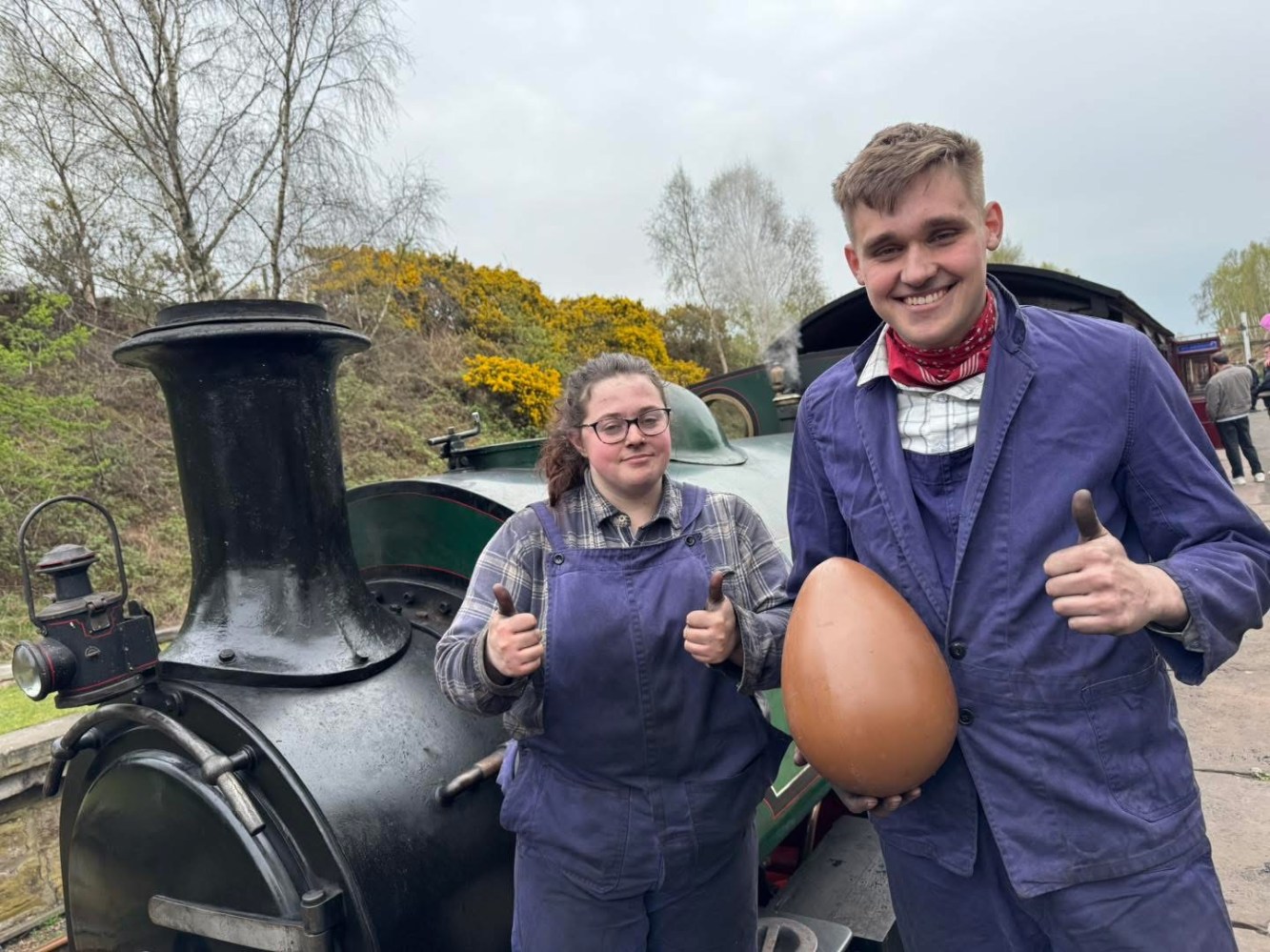 Two people in overalls give thumbs up near a steam train, one holds a large egg.