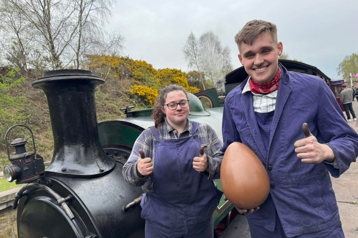 Two people in overalls give thumbs up near a steam train, one holds a large egg.