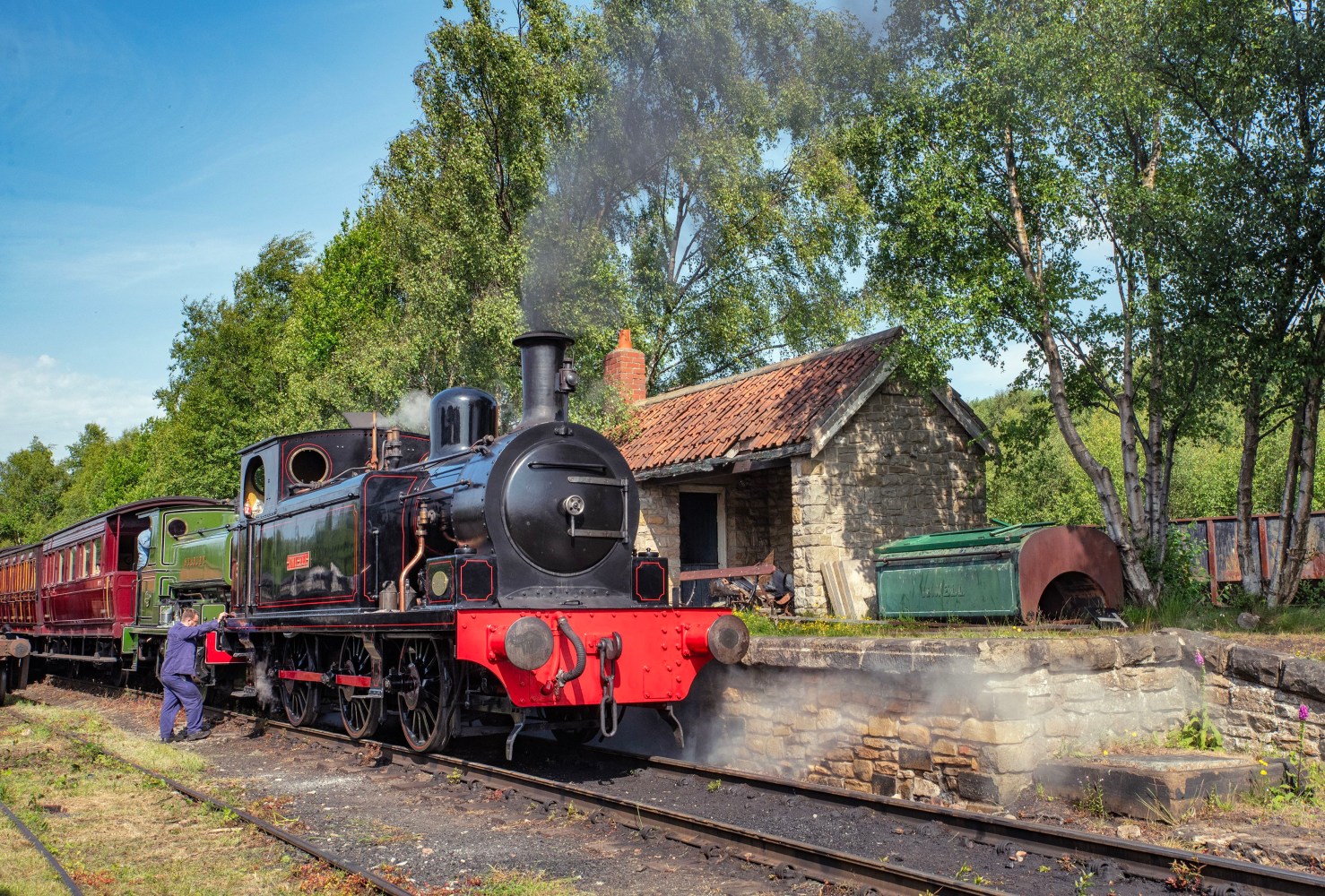 Vintage steam train at a rural station with trees and a stone building in the background.