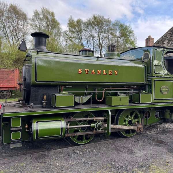 Green steam locomotive named 'STANLEY' parked outdoors near trees and a shed.