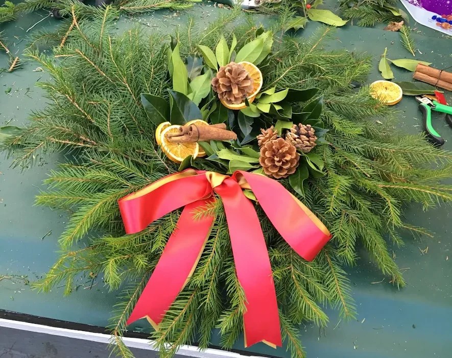 Festive wreath on table with pine, leaves, pinecones, orange slices, and red bow.
