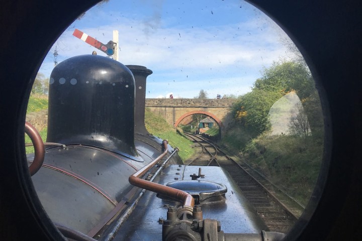 View through train window of steam engine with tracks and bridge ahead.