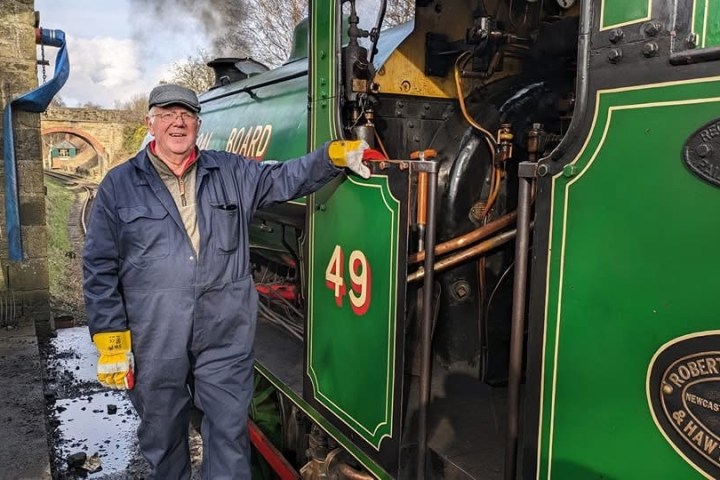Man in overalls stands beside a green steam locomotive.