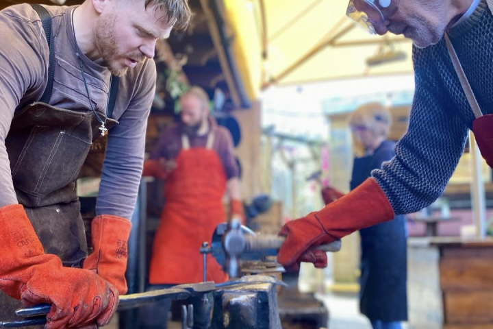 Two people in aprons forge metal on an anvil outdoors.