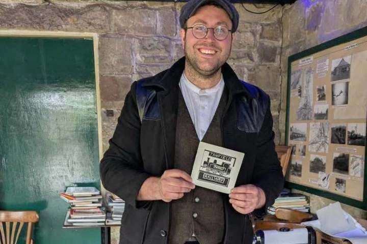 Man in vintage outfit holds a postcard inside a rustic room with books and photos.