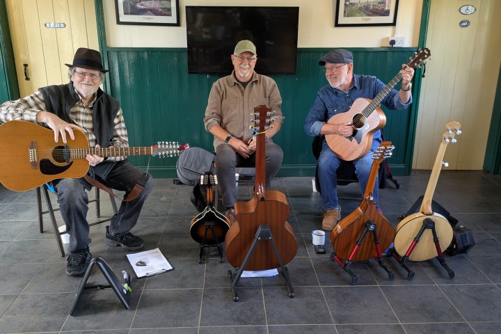 Three smiling musicians with guitars seated indoors, surrounded by string instruments.